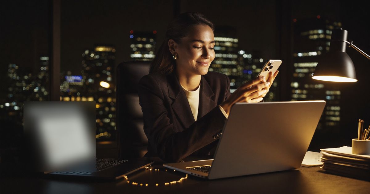 Une femme sourit en lisant un message sur son téléphone, le soir, dans un bureau face à la ville illuminée