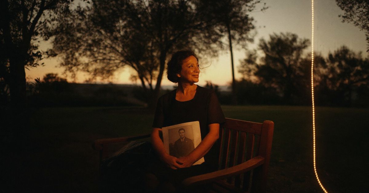Une femme souriante assise sur un banc parmi les oliviers au crépuscule, une photographie sur les genoux