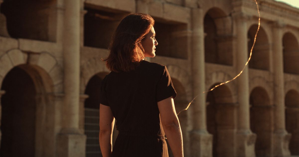Une femme marche le long des arcades des Arènes de Nîmes, dans la lumière dorée du soir