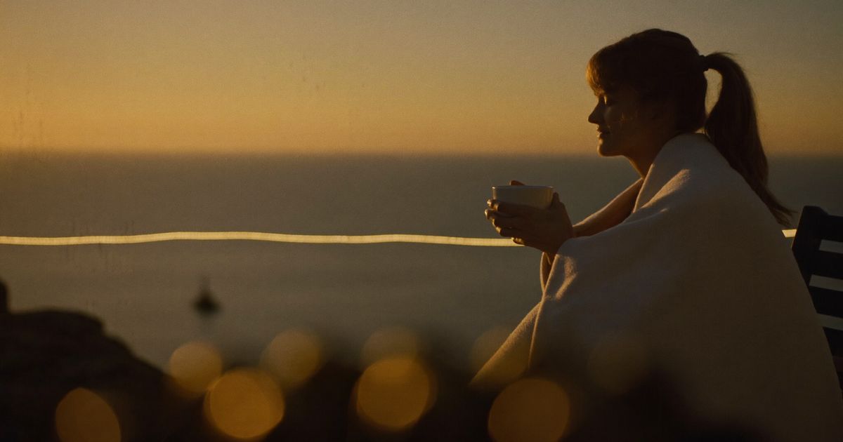 Une femme enveloppée dans une couverture, une tasse à la main, face à la mer au lever du soleil